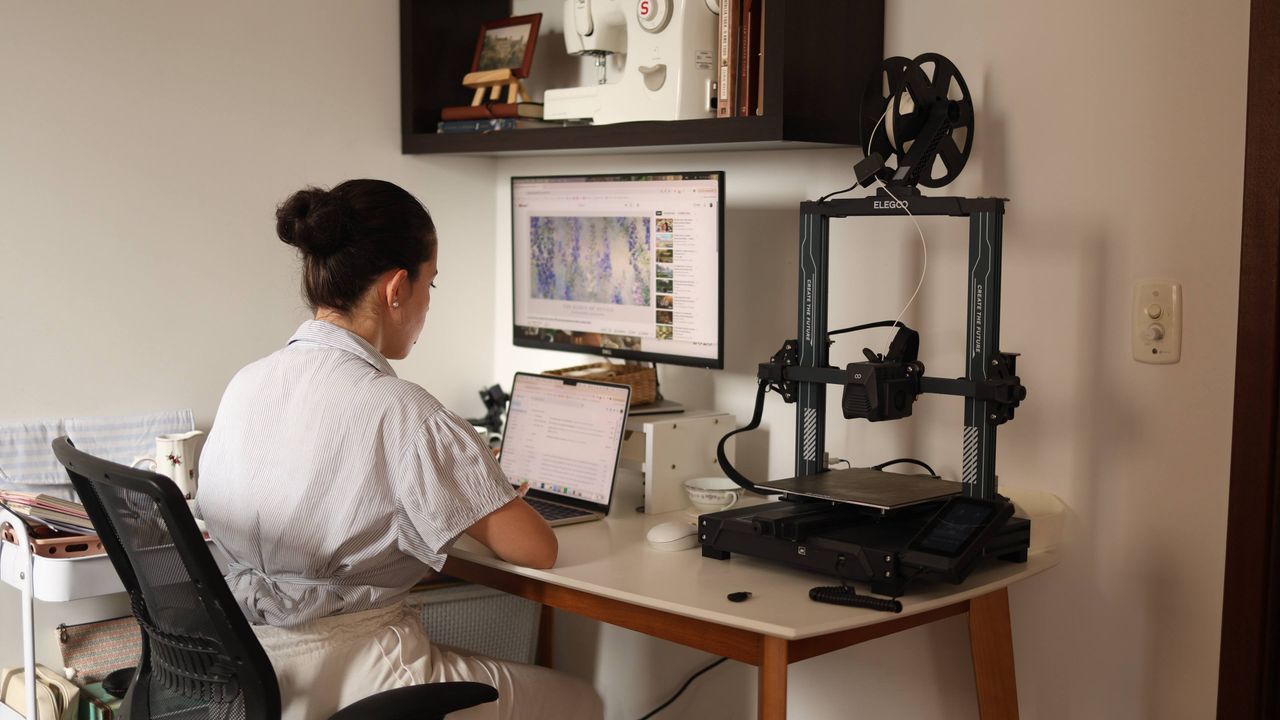 about-us A woman working on a laptop with a 3D printer in a modern home office setup.